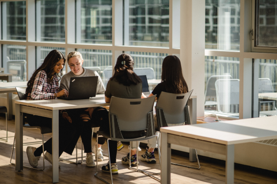 Four students sit at a table in the Kingston cafeteria. They share laptops and are smiling.